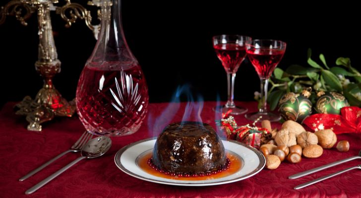 A christmas pudding with flaming brandy, a carafe and two glasses of wine, a spoon and fork, and a selection of nuts and Christmas baubles all sat upon a red velvet tablecloth