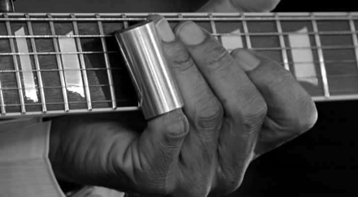 A hand playing a guitar with a metal cylinder covering one finger