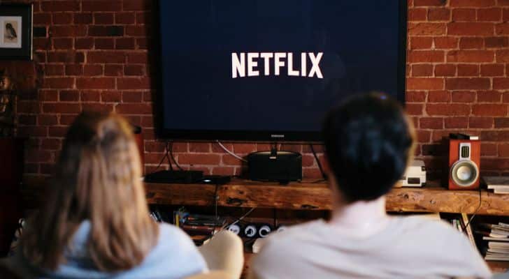 A couple sit on a sofa together watching a television in the background