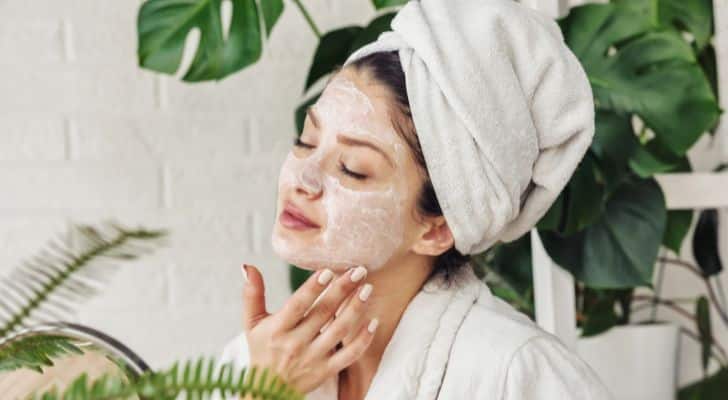 A woman in a white robe with a towel on her head applying skin cream to her face