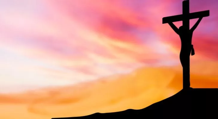 A person hanging off of a cross silhouetted against a colorful sky