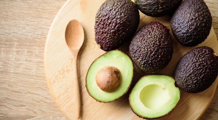 A selection of cut and uncut avocados on a chopping board next to a wooden spoon