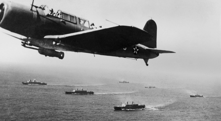 A WWII plane flies above a fleet of ships