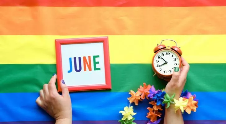 A person's hands holding a clock and a picture of the word "JUNE" on top of a pride flag