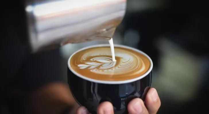 A barista holds a cup of fresh coffee and creates a pattern by pouring milk into it