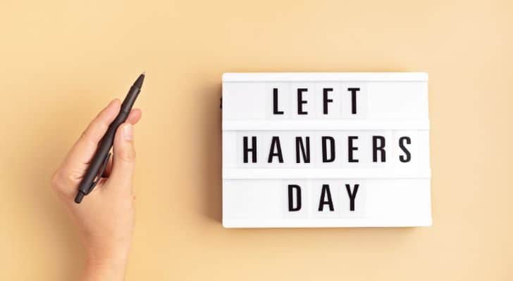 A person's left hand holding a pen next to a sign reading "Left hander day"