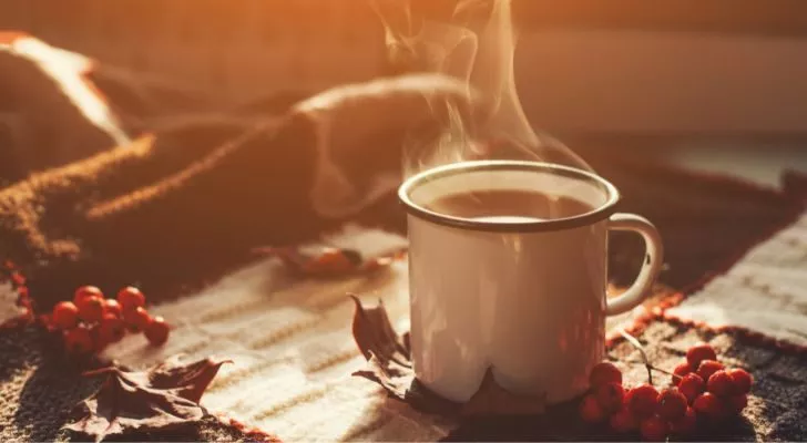 A steaming cup of coffee on a blanket surrounded by autumn leaves and spices