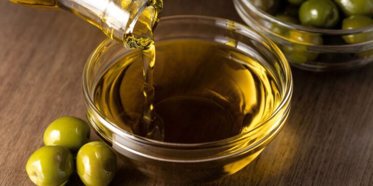 Olive oil pouring into a glass bowl, with green olives on the side.