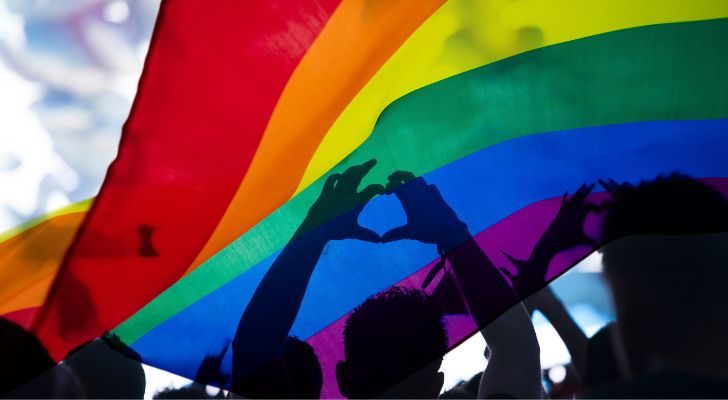 An LGBTQIA flag flies over a crowd as a man makes a heart shape with his hands