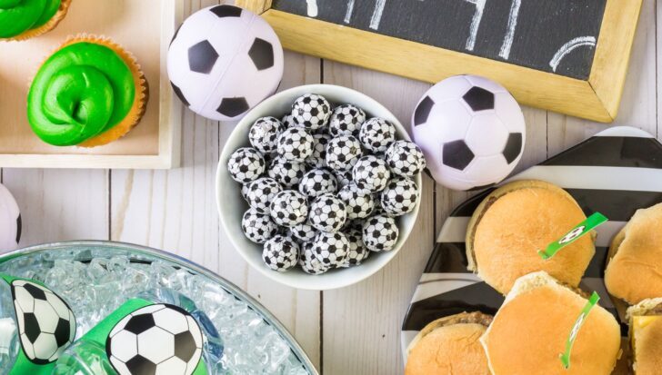 Soccer-themed party setup featuring green frosted cupcakes, soccer ball decorations, a bowl of small soccer ball chocolates, and sandwiches