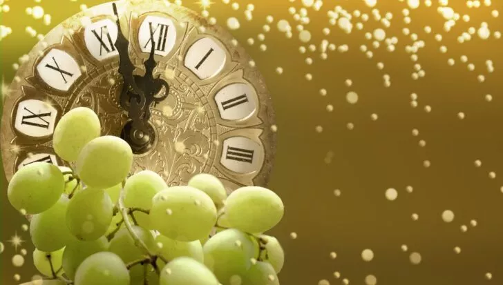 A bunch of green grapes in front of an ornate clock with Roman numerals, set against a gold sparkling background