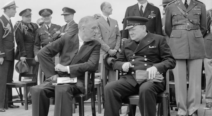 Winston Churchill and Roosevelt sitting next to each other surrounded by military personal