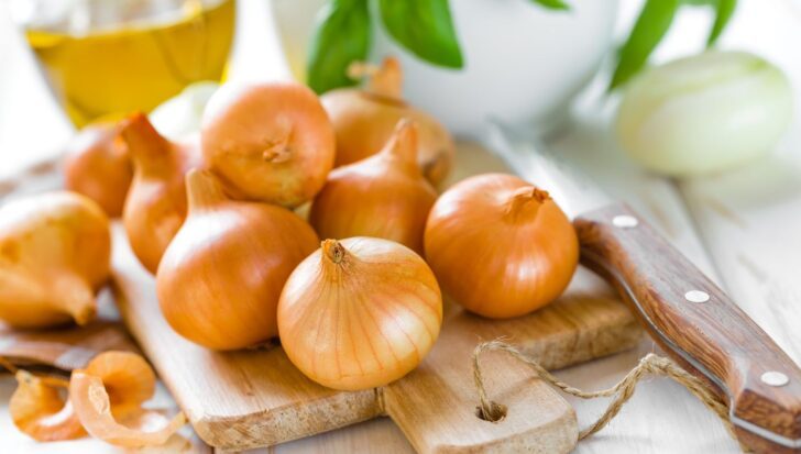 Several brown onions on a wooden cutting board with a knife nearby