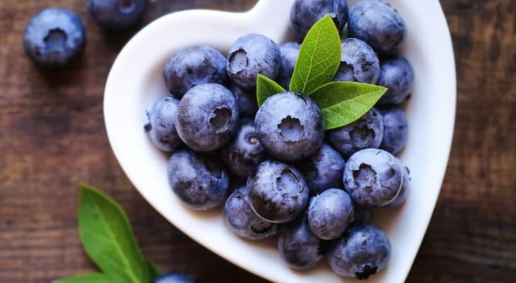 Some blueberries and green leaves arranged in a white, heart-shaped bowl