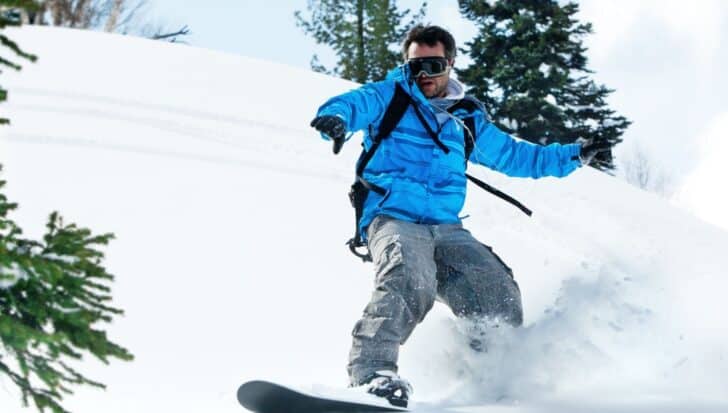 Person snowboarding down a snowy slope wearing a blue jacket and goggles, surrounded by trees