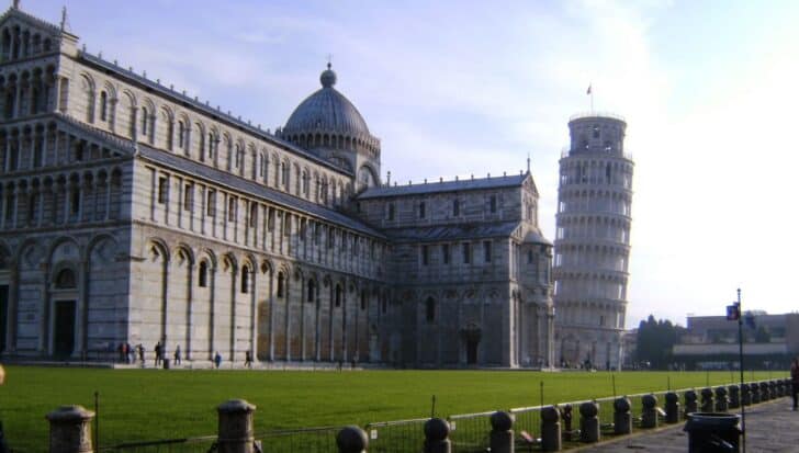 The Leaning Tower of Pisa next to the Pisa Cathedral on a sunny day with a green lawn in the foreground and a few people walking