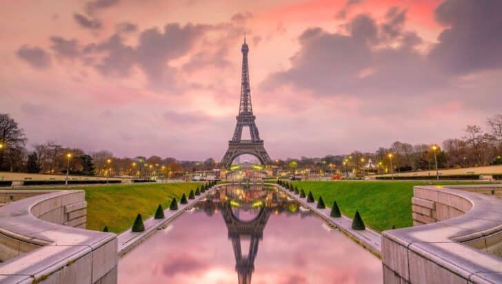 The Eiffel Tower at sunset with a reflection in a pond, surrounded by gardens and trees