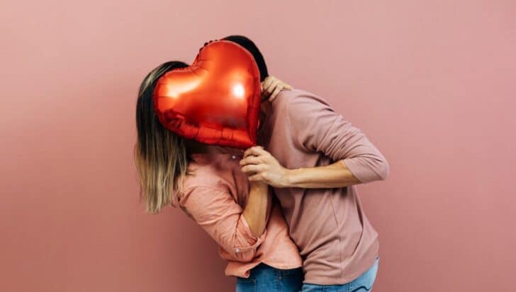 Two people holding a red heart-shaped balloon in front of their faces in a playful gesture, both wearing pink tops against a pink background